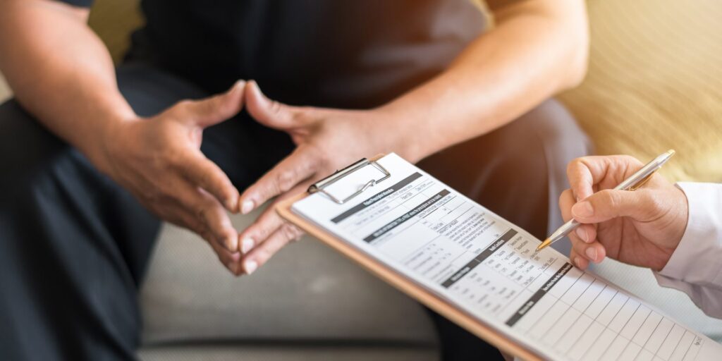 A man wearing dark clothes sitting down at a doctor's office and a physician takes notes of what he is saying. Representing how one can benefit from calling a Portland criminal defense attorney.