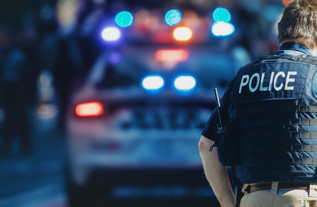 A police officer walking, and a patrol car with lights on in the background, representing how one can benefit from calling a Portland criminal defense attorney.