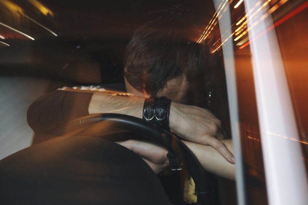 An upset man with burying his face between his arms and the steering wheel of a car. Representing how one can benefit from calling a Portland criminal defense attorney.