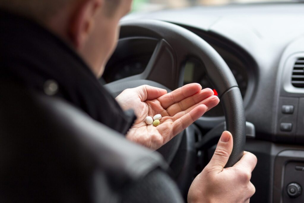 A man wearing a dark coat, sitting on the driver's side of a car taking some pills. Representing how one can benefit from calling a Portland criminal defense lawyer.
