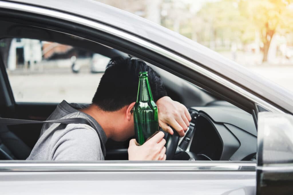 An intoxicated man in the driver seat of a car holding a bottle, representing how one can benefit from calling a Portland criminal defense lawyer.
