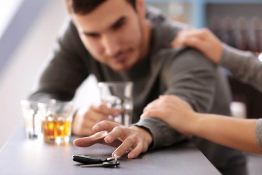 An intoxicated man holding a glass of alcohol reaching out for a set of car keys while somebody else is holding him back, representing how one can benefit from calling a Portland criminal defense lawyer.