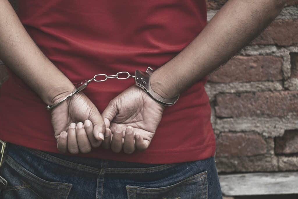 A man wearing a red shirt with his hands behind his back wearing handcuffs, representing how one can benefit from calling a Portland criminal defense lawyer.