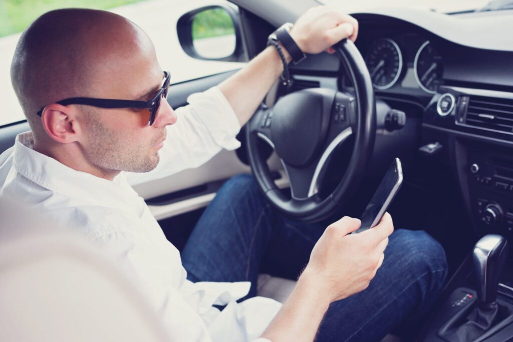 A man inside a car, sitting on the driver's seat holding the wheel with one hand and his phone with the other, representing how one can benefit from calling a Portland criminal defense lawyer.