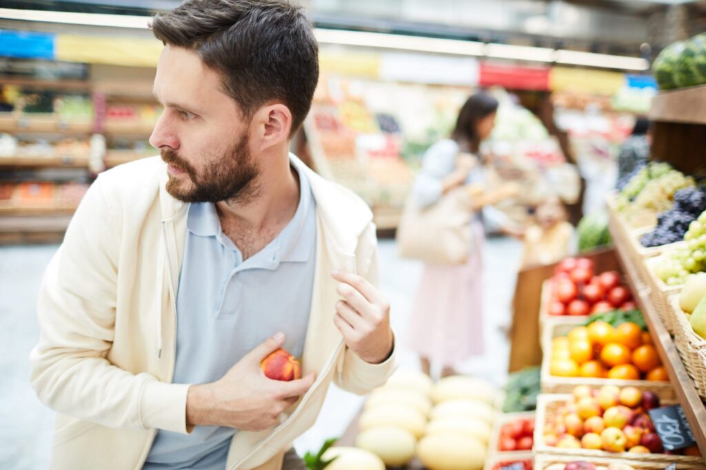 A man in a grocery store looking around while he puts a piece of fruit inside his sweater, representing how one can benefit from calling a Portland criminal defense lawyer.