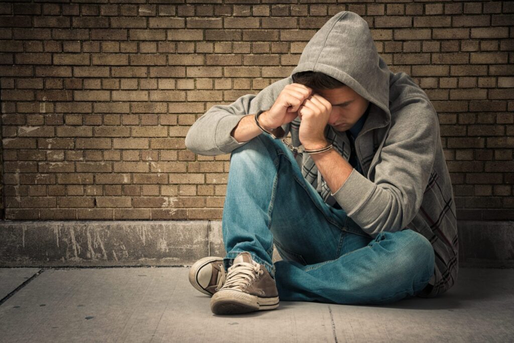 A teenager wearing a hoodie is against a wall wearing handcuffs, representing how one can benefit from calling a Portland criminal defense lawyer.
