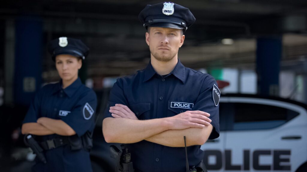 Two police officers looking straight with a police car in the background, representing how one can benefit from calling a Portland criminal defense attorney.