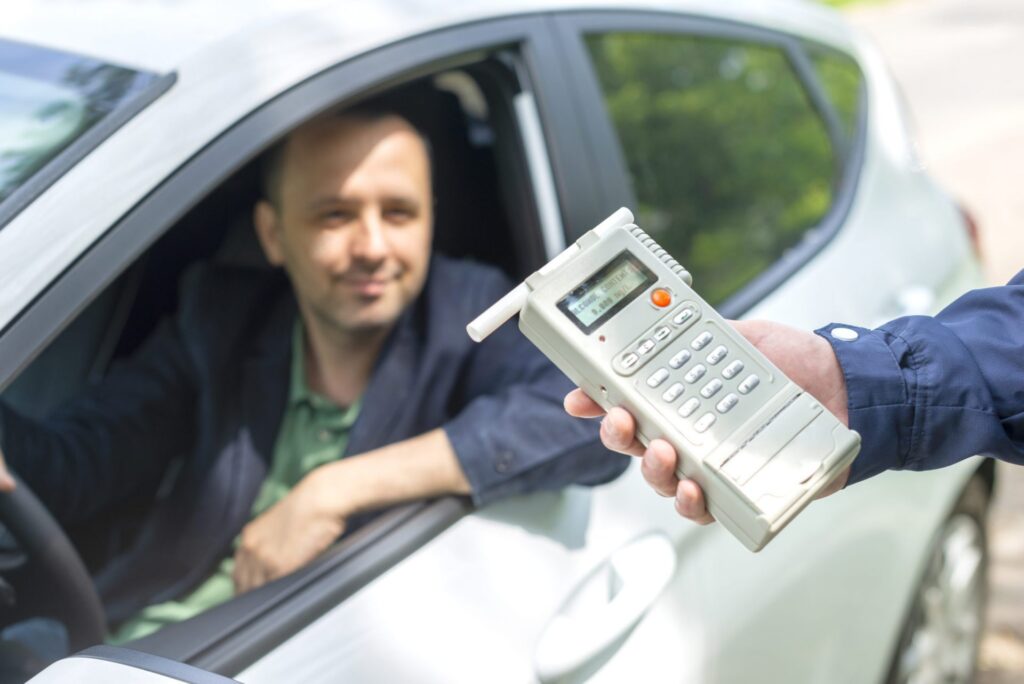 A cop approaching a man in the driver seat of a car with a breathlyzer, representing how one can benefit from calling a Maine criminal defense lawyer