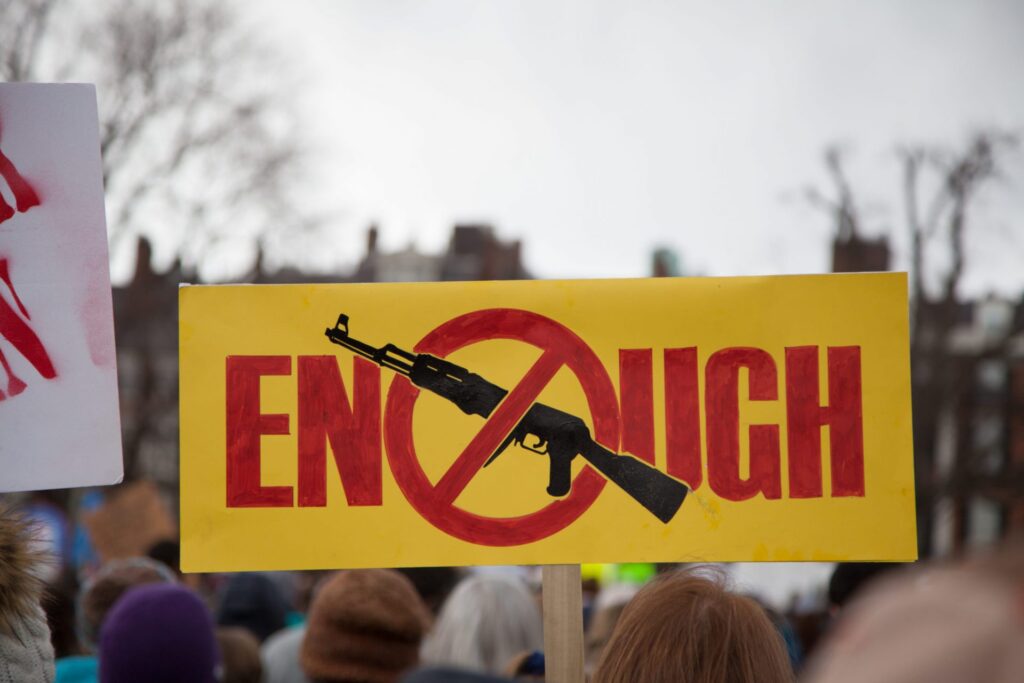 People in a march holding a yellow sign that says "Enough" and picture of a shotgun being banned, representing how one can benefit from calling a Maine criminal defense lawyer.